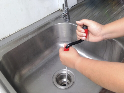 A woman using a tool to break up a clog in the kitchen sink.